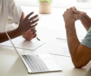 Two people sit at a desk with a laptop and documents, having a discussion in a bright office setting.