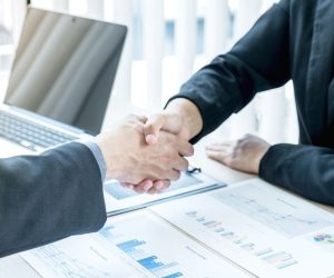 Two people in business attire shake hands over a desk with charts and a laptop.