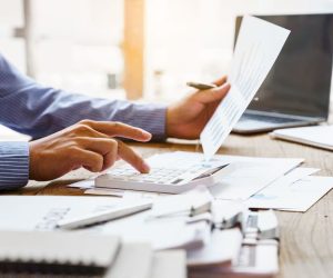 Person using a calculator and holding a financial document at a desk with papers and a laptop.