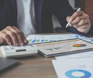 Person using a calculator and tablet with charts and graphs on a desk covered in financial documents and reports.
