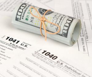 A rolled stack of $100 bills tied with string sits on U.S. tax forms 1040 and 1041.